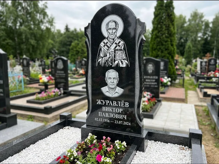 Памятник с гравировкой иконы: стела с образом в полный рост на обороте — гравировка образа Матроны Московской, низкие кусты спиреи в цвету, работа «Лебедева» в Южно-Сахалинске